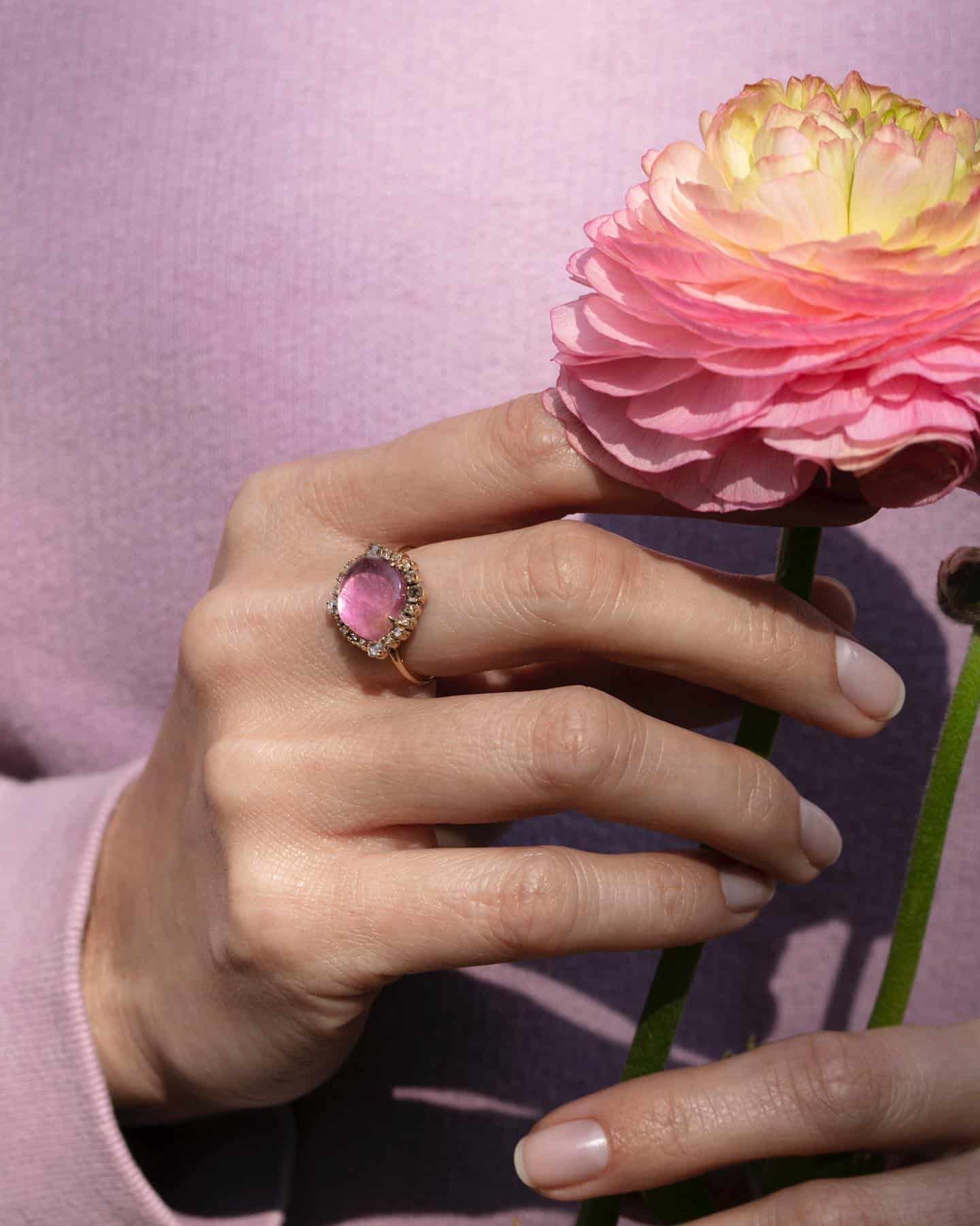 Main portant une bague rose tenant une fleur rose et jaune sur un fond rose.