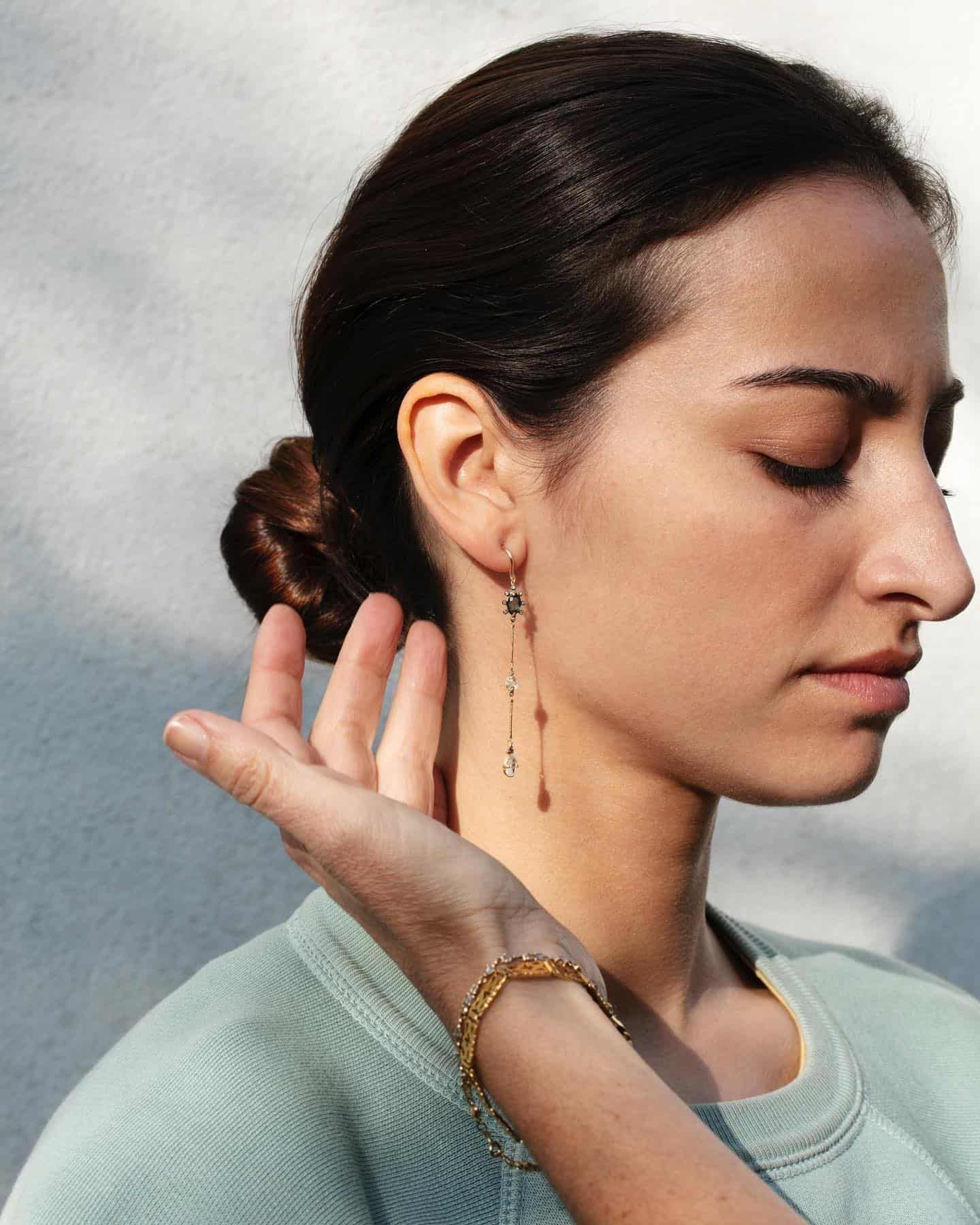 Femme aux cheveux noirs, portant des boucles d'oreilles et un bracelet en or, la main levée, les yeux fermés.