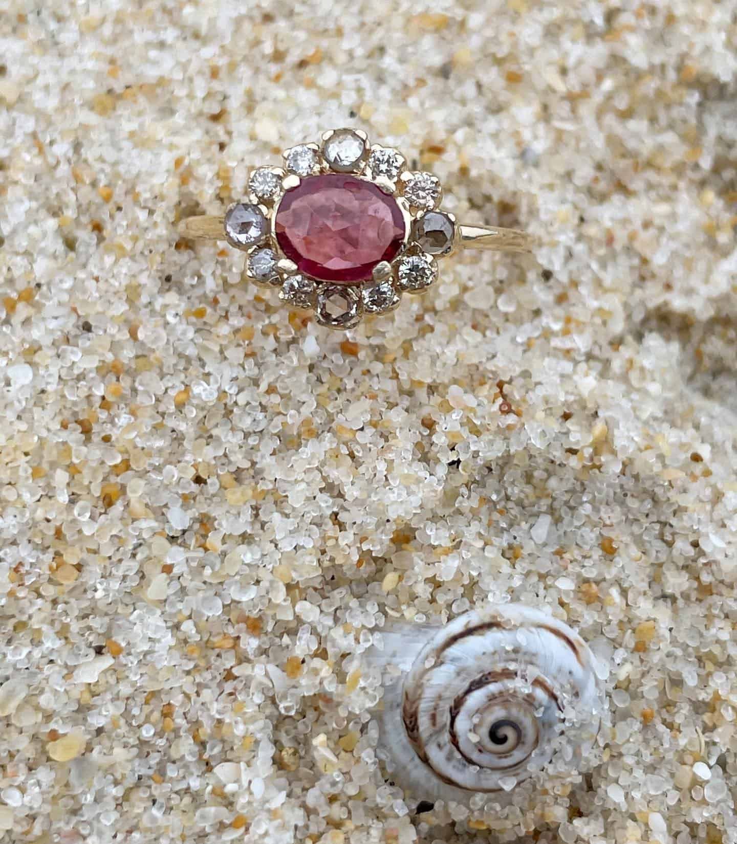 Bague en or avec pierre rouge et diamants environnants sur du sable près d'un coquillage.