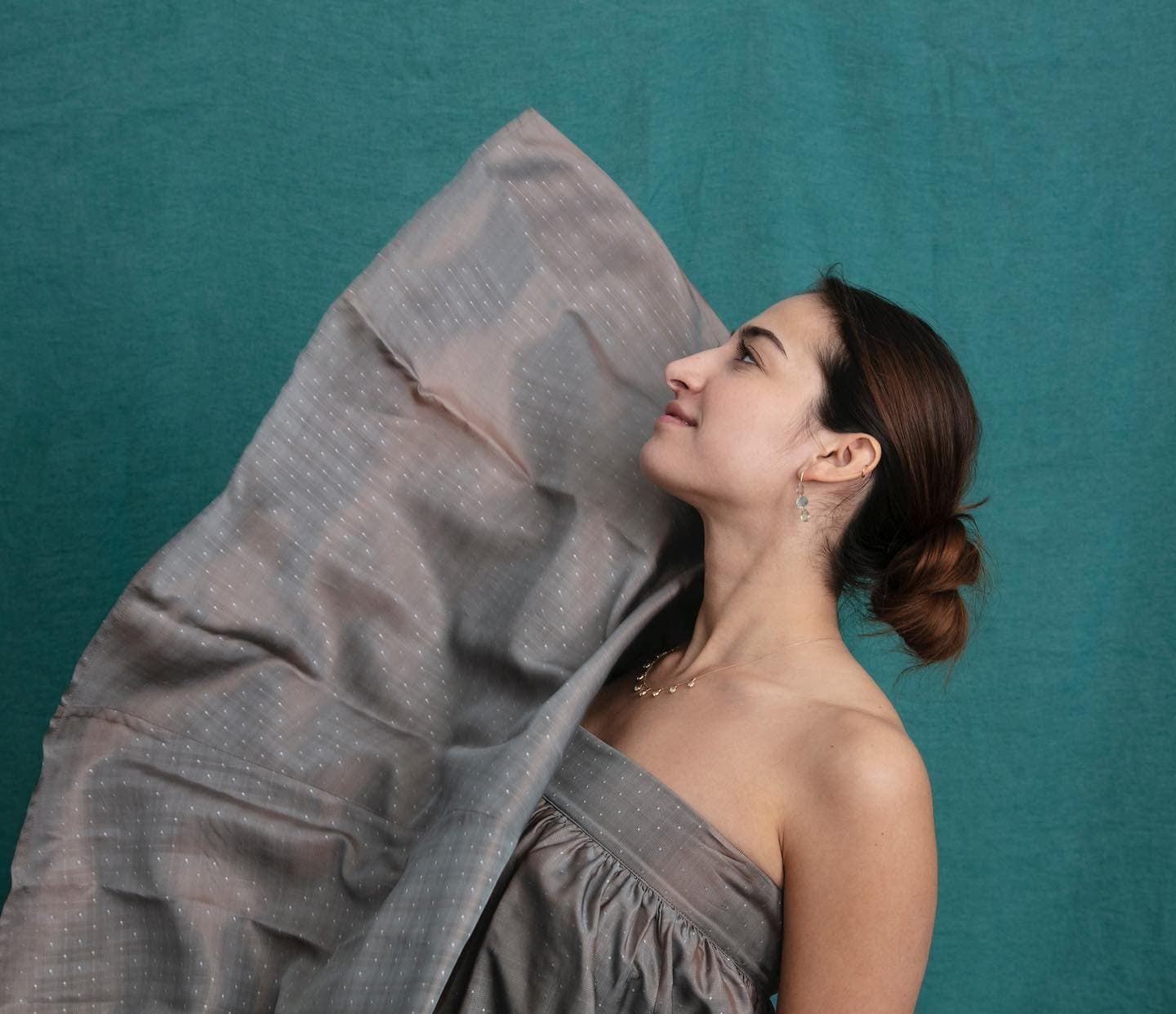Une femme en robe grise lève les yeux, tenant un tissu sur fond bleu sarcelle.