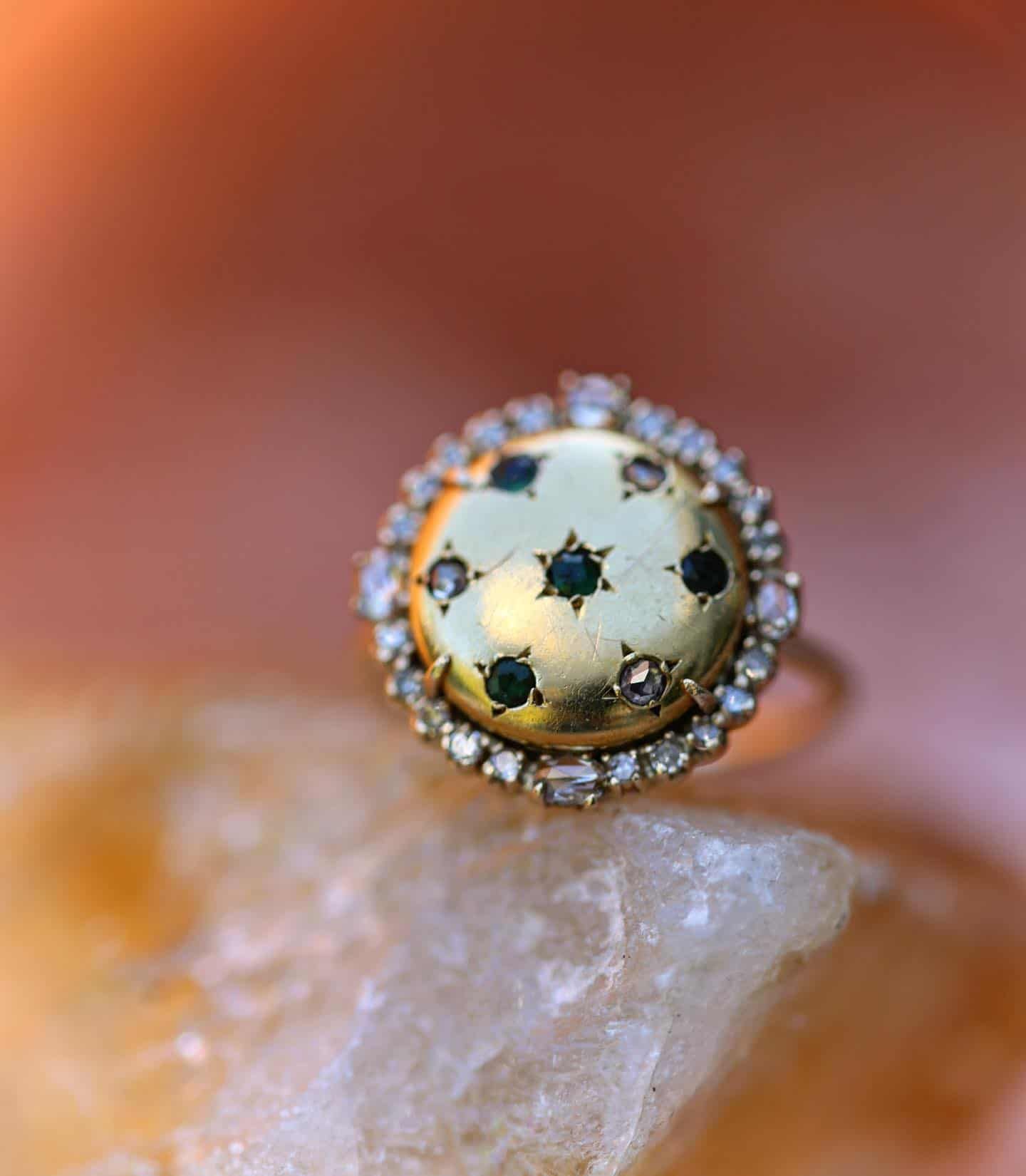 Bague en or avec un visage circulaire, détails en émeraude et diamant, reposant sur une pierre de couleur claire.