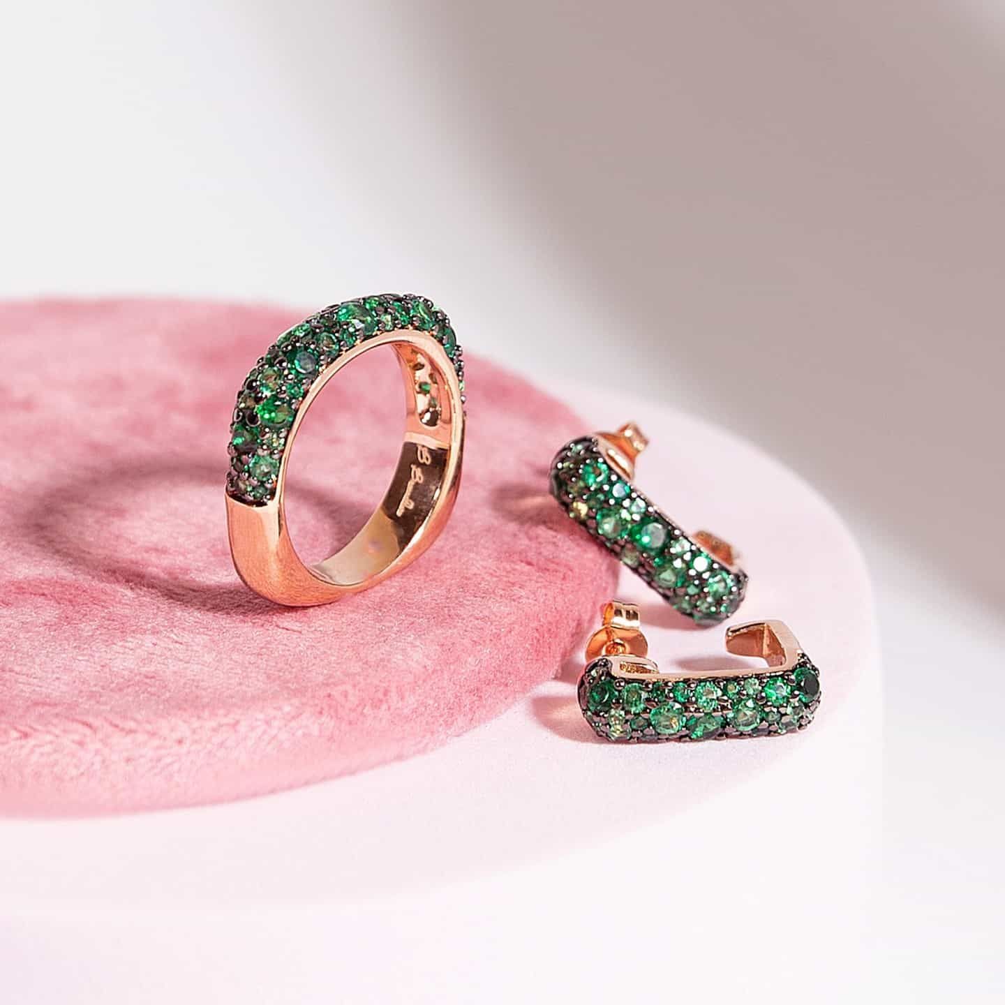 Bague et boucles d'oreilles en or rose serties de pierres précieuses vert émeraude sur une surface rose.