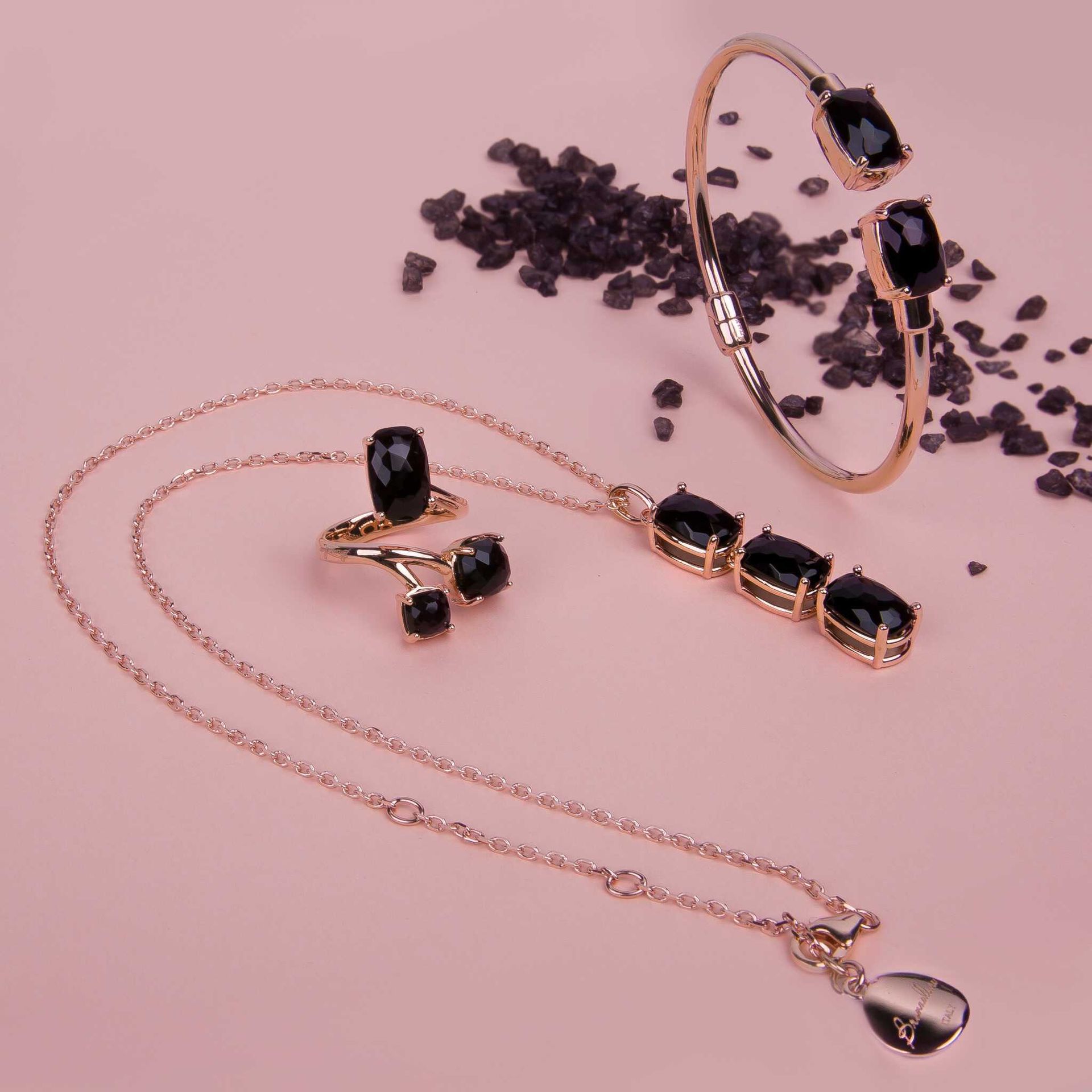 Ensemble de bijoux avec chaîne, bague et bracelet en or rose avec des accents de pierres précieuses noires, sur une surface rose avec des granules noirs.