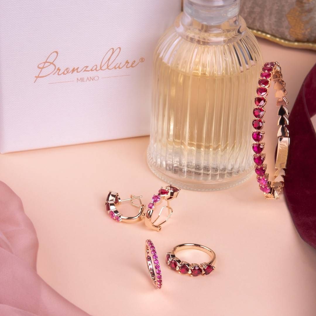 Ensemble de bijoux avec des accents dorés et des pierres précieuses roses disposées avec du parfum sur une surface pêche.