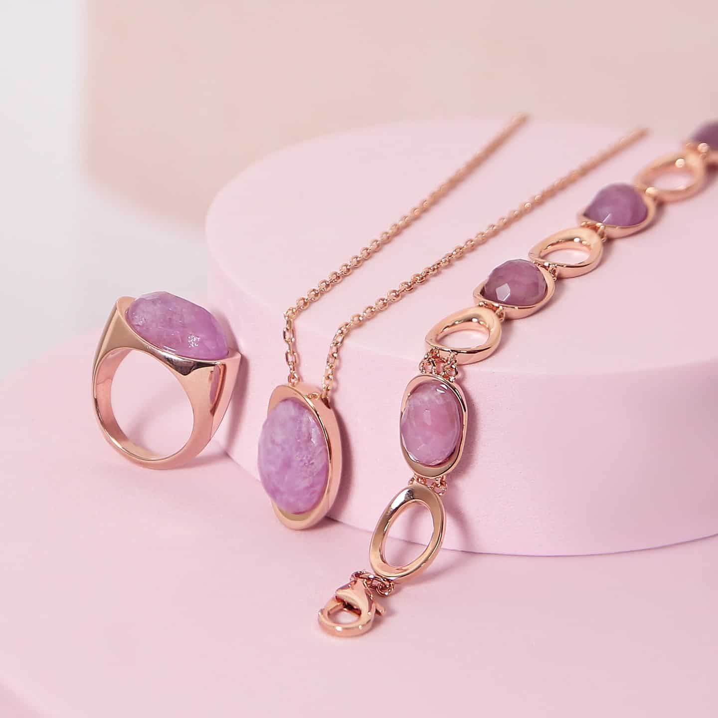 Parure de bijoux en or rose : bague, collier et bracelet, avec pierres précieuses roses sur fond rose.