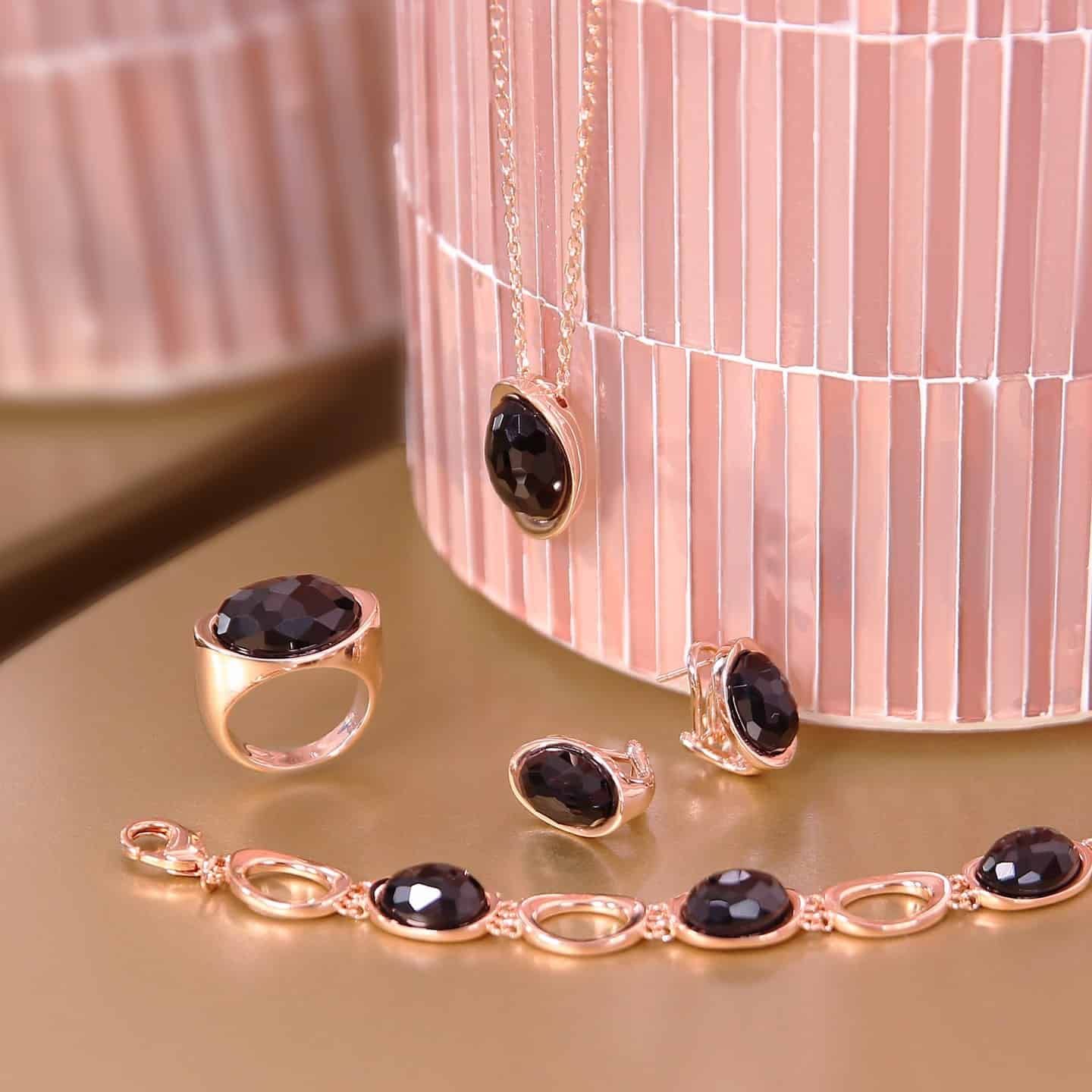 Parure de bijoux en or : collier, bague, boucles d'oreilles, bracelet, avec pierres noires. Fond rose.