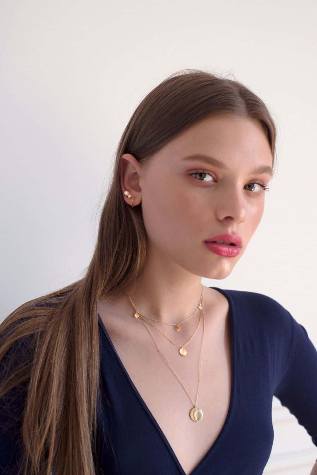 Femme aux longs cheveux bruns portant des bijoux en or et un haut bleu, regardant la caméra.