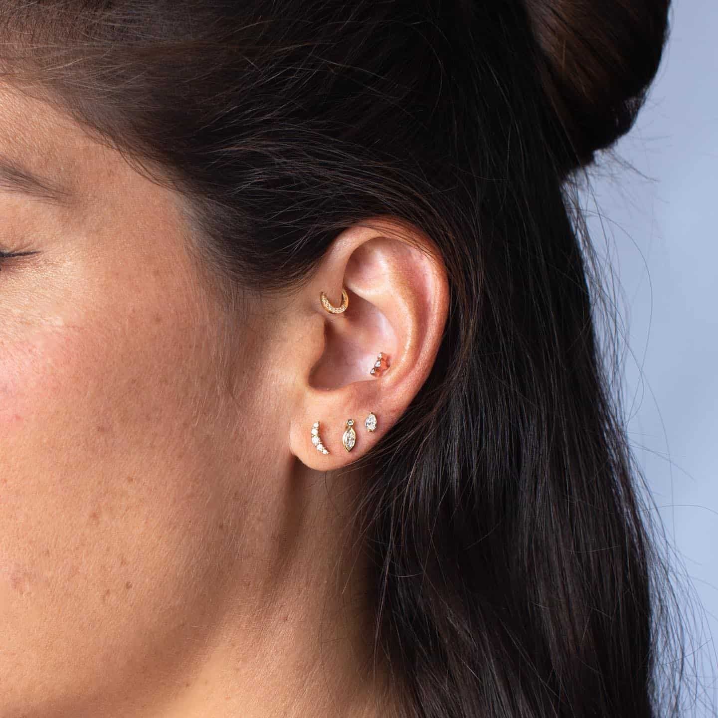 Oreille de femme avec plusieurs boucles d'oreilles en or, dont des créoles et des clous, sur fond bleu.