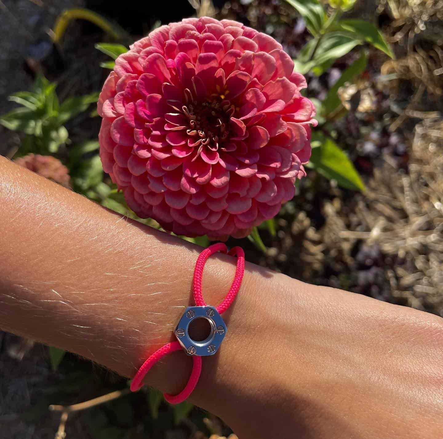 Un poignet portant un bracelet en cordon rose avec une breloque en forme de noix en argent ; une fleur de zinnia rose en arrière-plan.