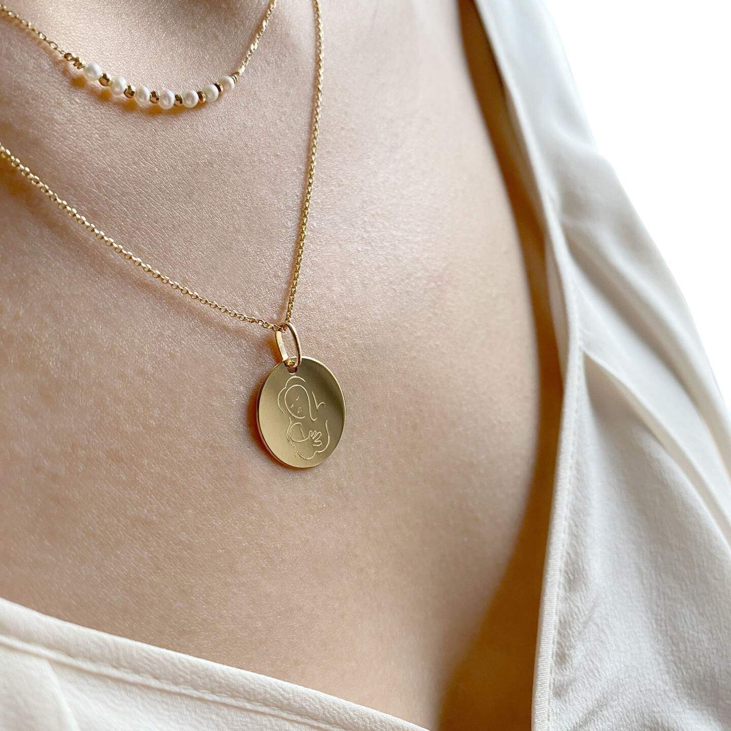 Une femme portant deux colliers en or, l'un avec un pendentif, sur un haut de couleur neutre.