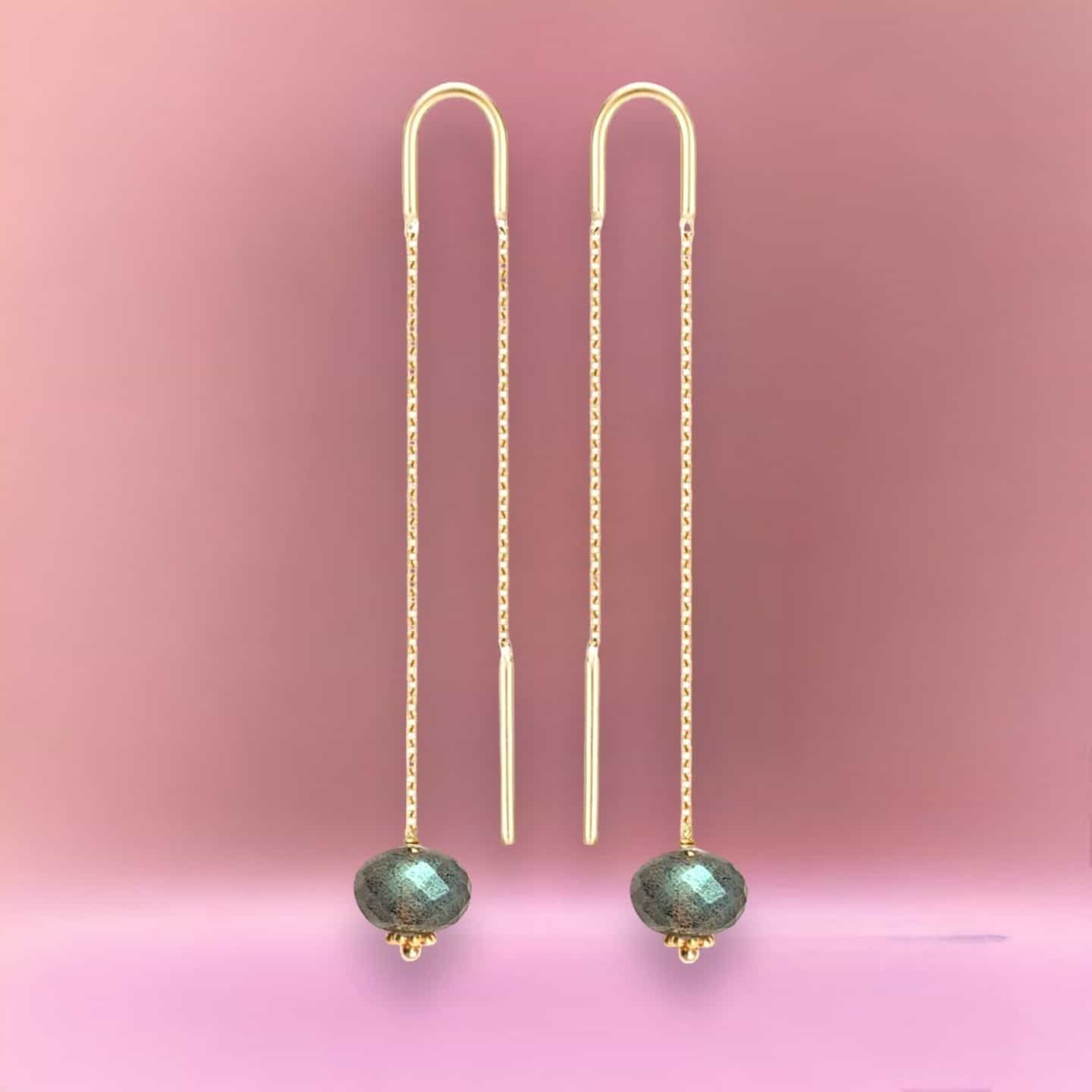 Boucles d'oreilles enfileuses en or avec perles facettées vertes sur fond rose.