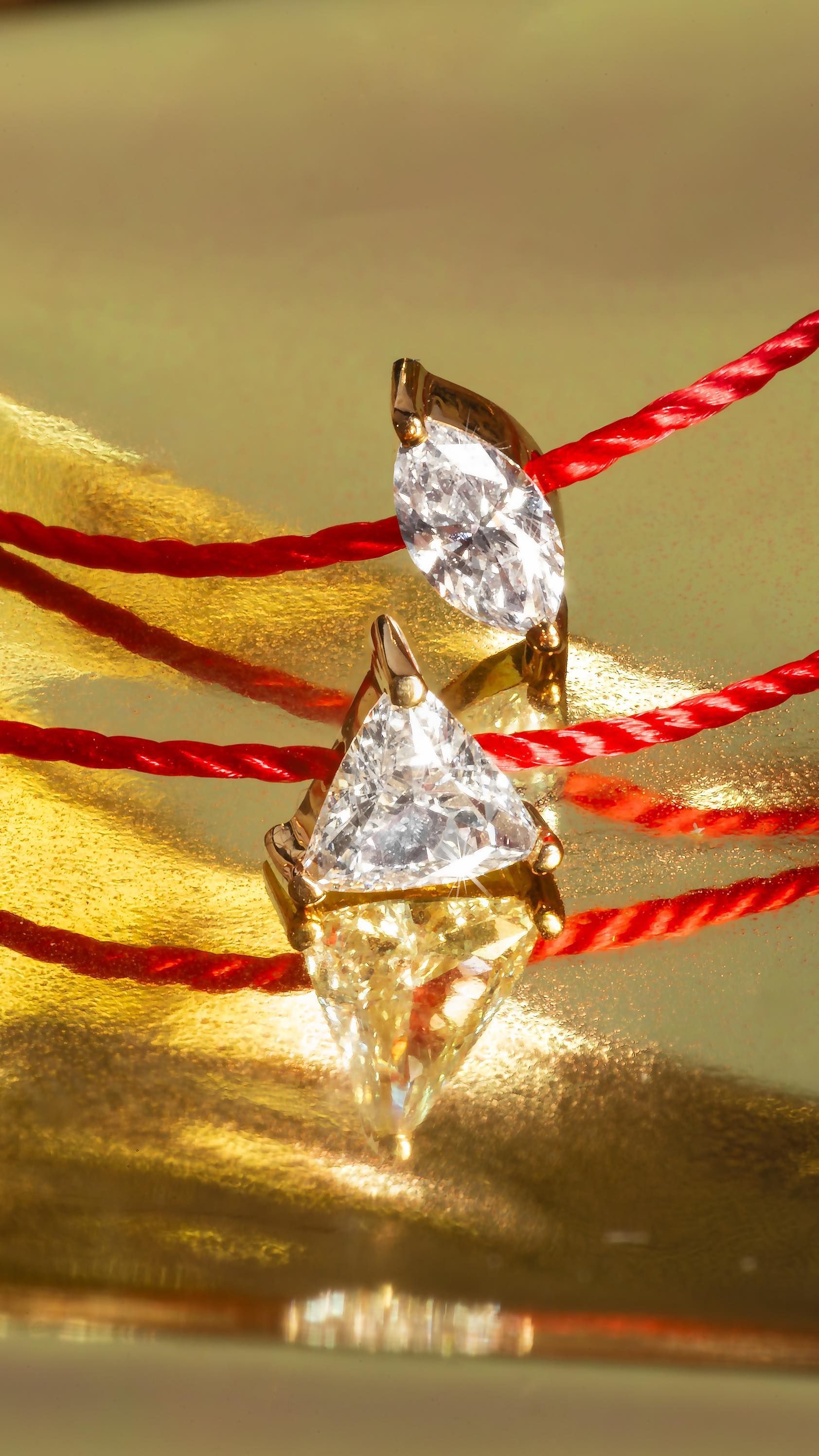 Deux breloques en or en forme de diamant sur des fils rouges, se reflétant sur une surface dorée.