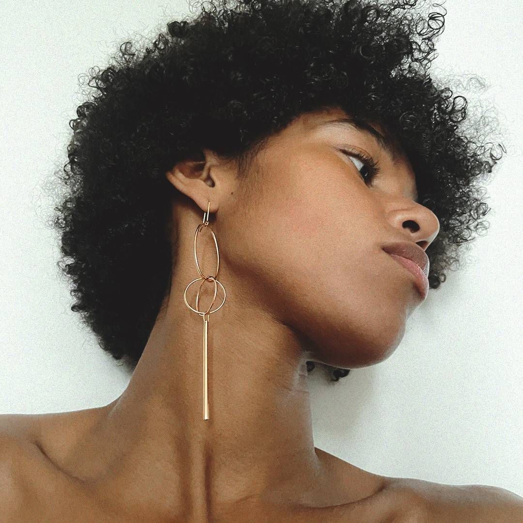 Femme aux cheveux noirs bouclés portant une longue boucle d'oreille en or. Elle regarde vers sa droite avec une expression sérieuse.