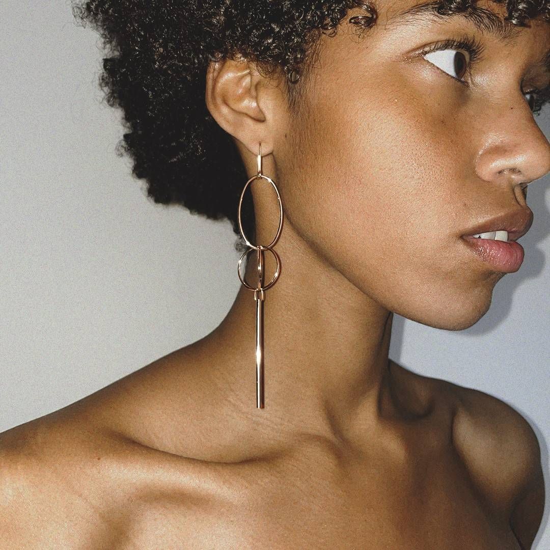Femme aux cheveux bouclés portant de longues boucles d'oreilles en or, regardant hors caméra.
