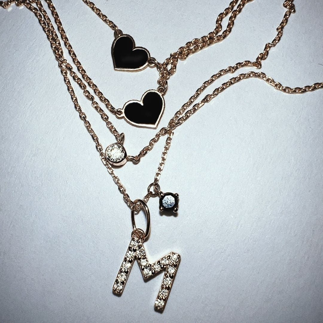 Colliers superposés en or rose avec pendentifs en forme de cœur noir, une breloque « M » et un diamant.