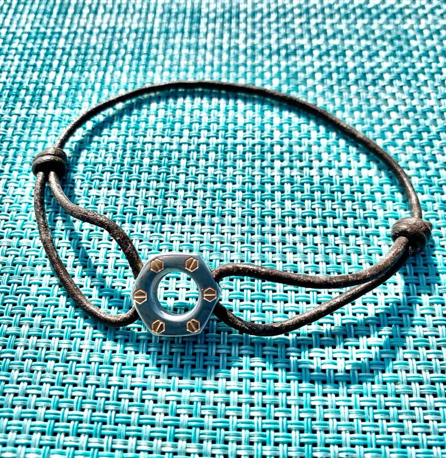 Bracelet cordon noir avec une breloque hexagonale en argent et or sur une surface tissée turquoise.