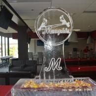 Clear ice sculpture with “M” logo and decorative table display in a banquet hall
