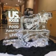 US Foods ice sculpture on a table at an indoor event, with a black cloth backdrop and branding signage behind it