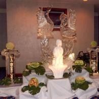 White statue with ice sculptured swans in the background with flowers, candles, and draped cloth in a dim room