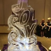 Clear ice sculpture spelling “JOY” glowing at an event, with people standing in the background.