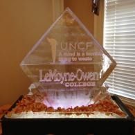 Clear ice sculptured award with etched logos on a table in front of window blinds
