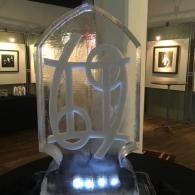 Clear ice sculpture lit from below in a museum-like gallery, with framed artworks on the walls.
