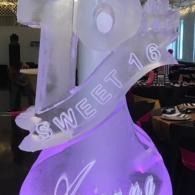 Glowing translucent ice sculpture with “SWEET 16” engraved, lit purple at a banquet table.