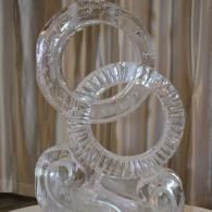 Clear acrylic interlocking rings sculpture on a pedestal, with beige curtains in the background