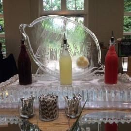 A bar display with a clear etched sign, three colorful bottles, and rows of glasses on a counter.