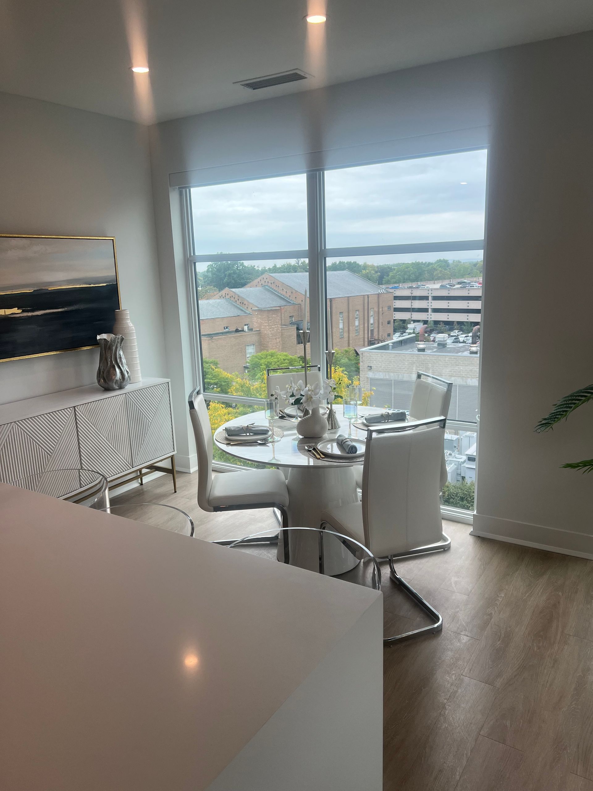 Dining area with a round table, white chairs, and a large window overlooking a city.