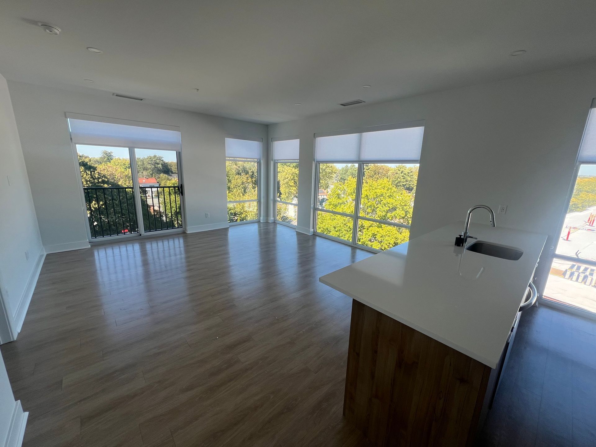 Empty living space with large windows overlooking trees; wood floors and a kitchen island.