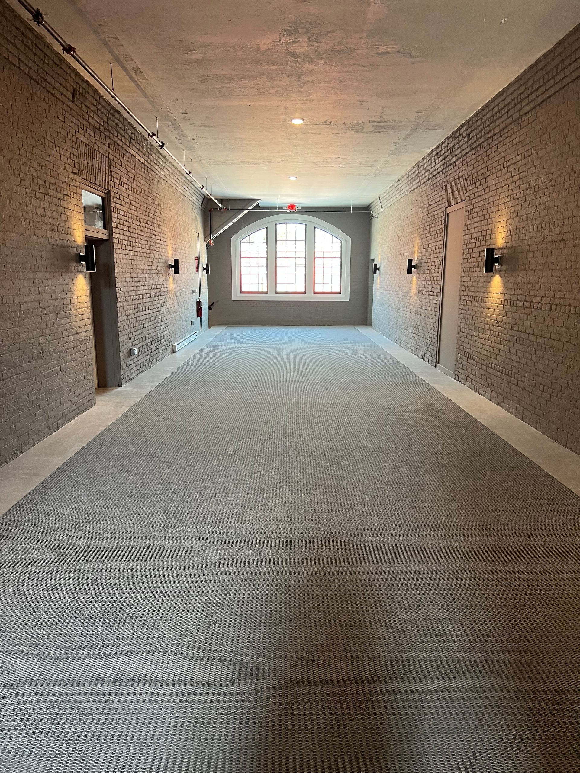 Hallway with patterned carpet, brick walls, sconce lighting, and a window at the end.