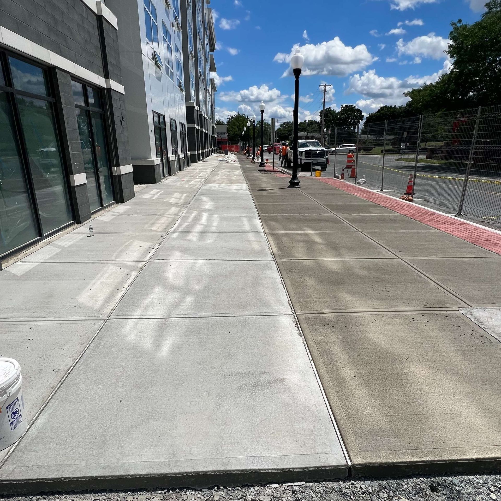 Sidewalk being cleaned; half gray, half light. Construction fence and building in the background. Bright day.