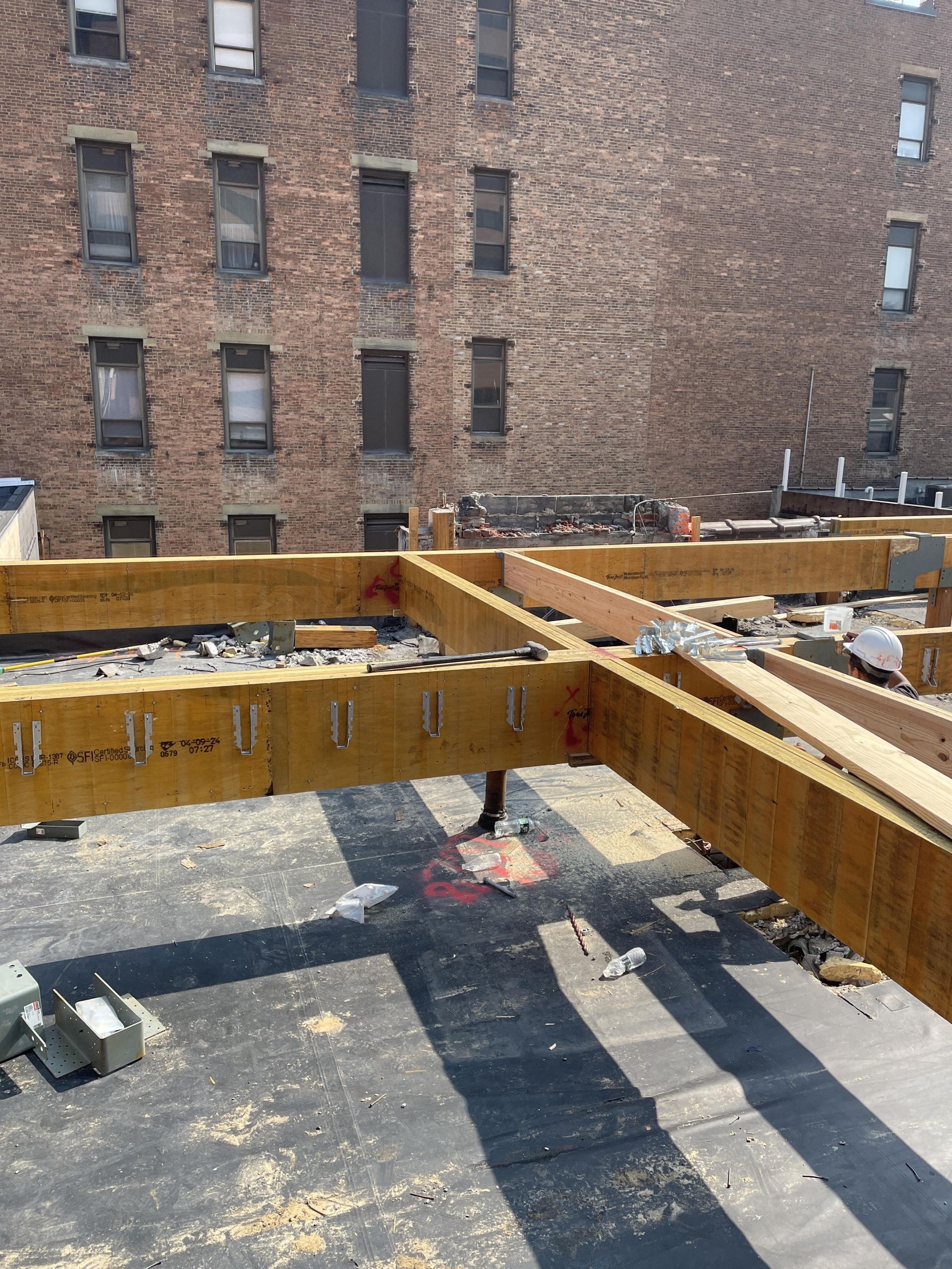 Rooftop construction with wooden beams, black roofing material, and a brick building backdrop.