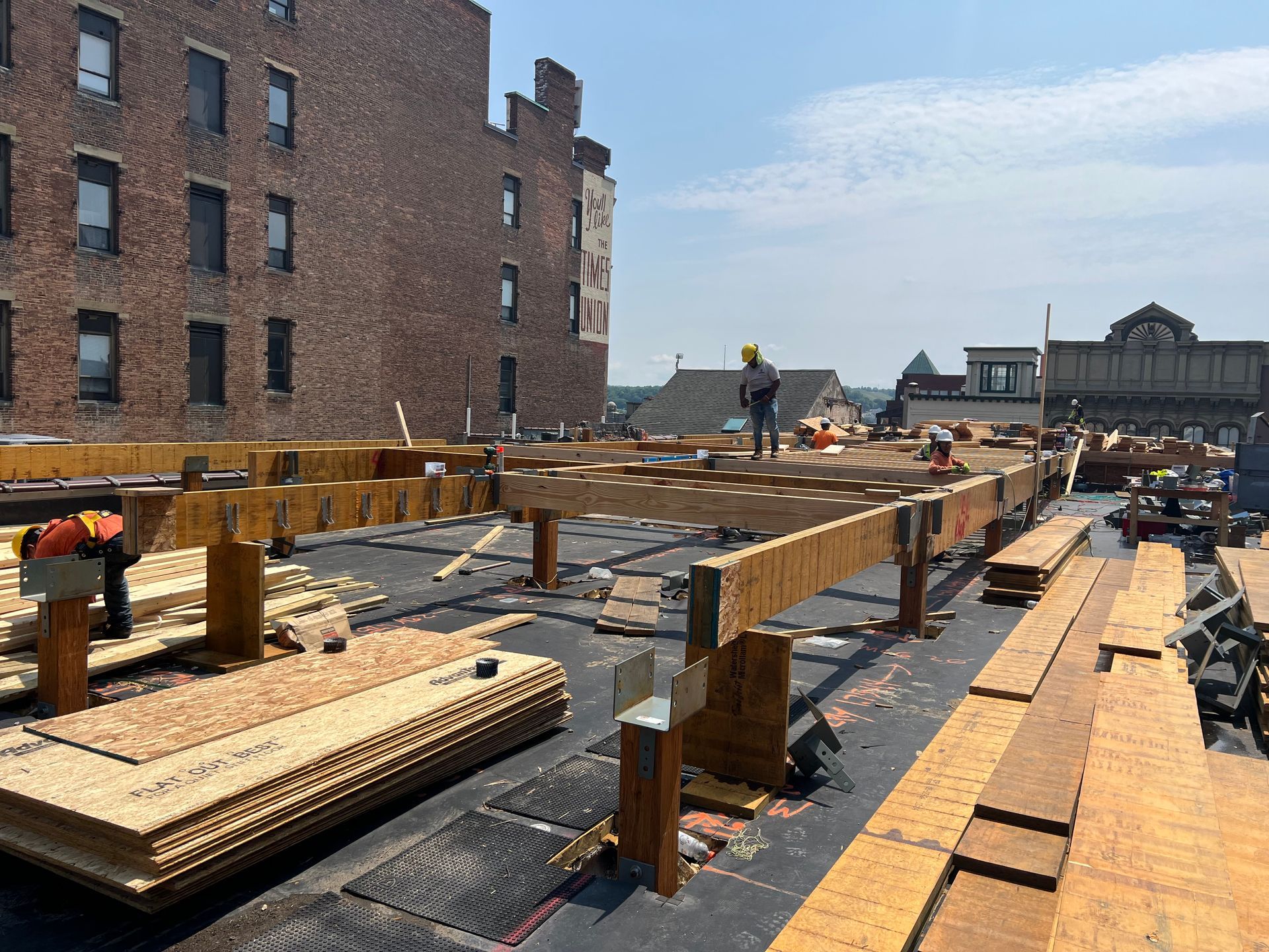 Construction workers on a rooftop, building a wooden structure. Bricks, blue sky, and other buildings are visible.