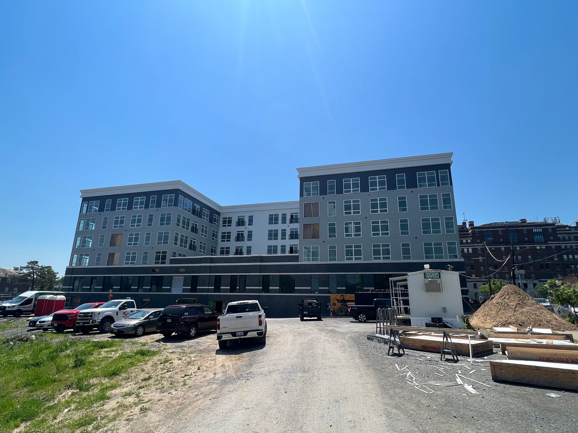 Multi-story gray apartment building with a flat roof under a bright blue sky; vehicles parked in front.