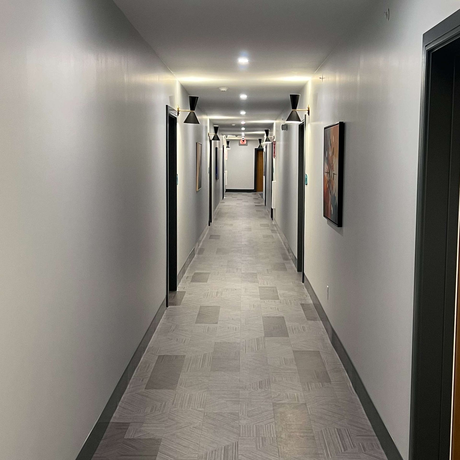 Long, narrow hallway with doors on both sides; gray walls, tiled floor, and ceiling lights.