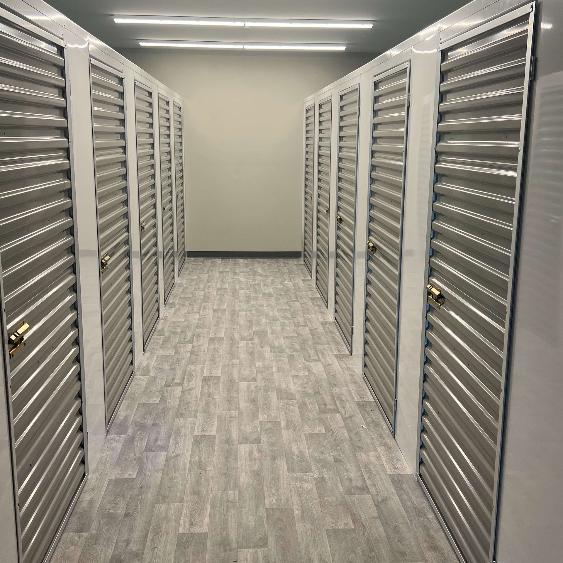 Rows of closed storage unit doors in a well-lit hallway with light grey flooring.