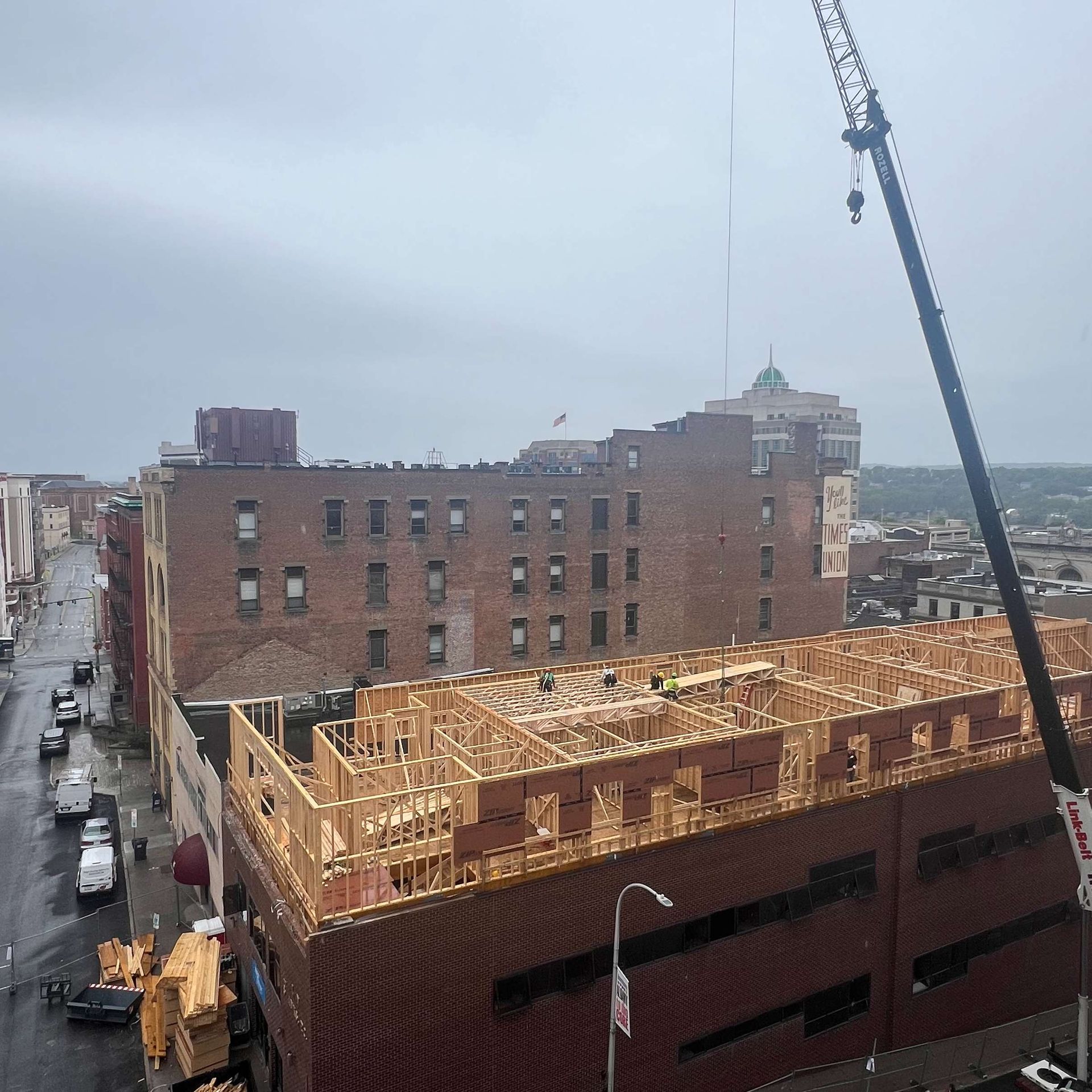 Construction of a building with a crane; workers on the wooden frame. Street with vehicles in the background. Overcast day.
