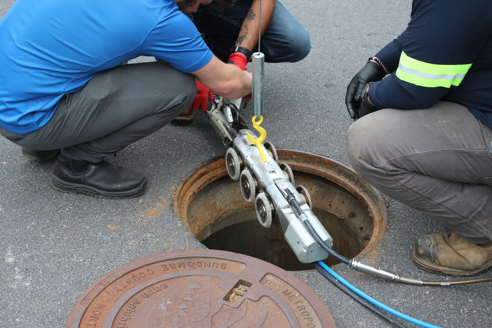 Two workers use a CCTV robot to inspect a manhole opening on asphalt.