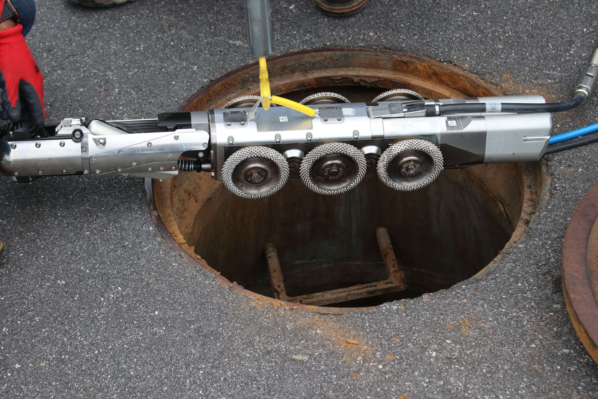 Metal CCTV robotic device being lowered into an open manhole with a yellow strap attached
