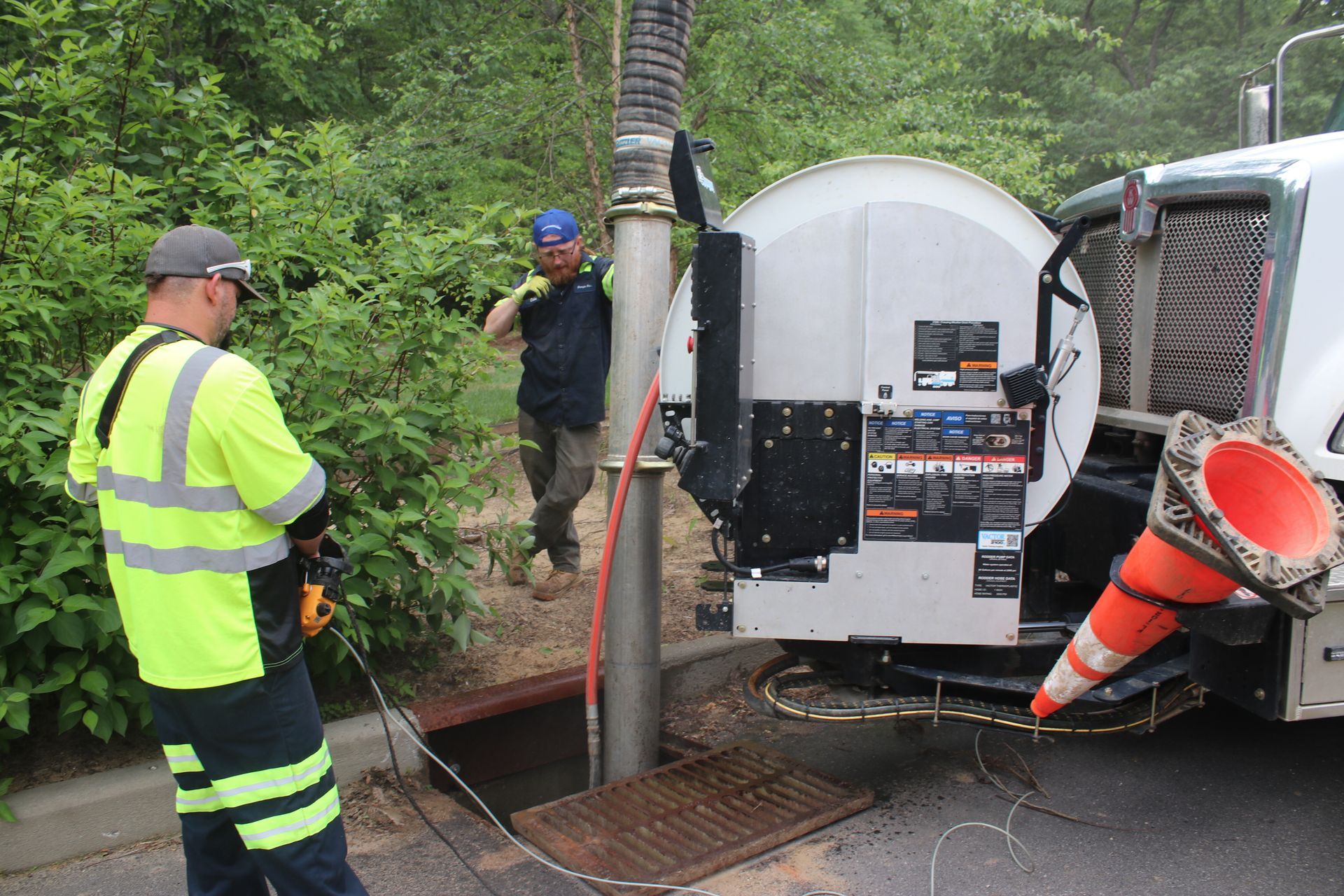 Workers operating a sewer cleaning truck beside an open manhole on a roadside in a wooded area. Vac truck in Asheville