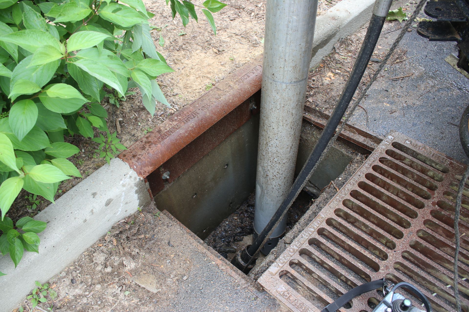 Open sidewalk drain with metal grating, concrete curb, and a vertical pipe beside greenery