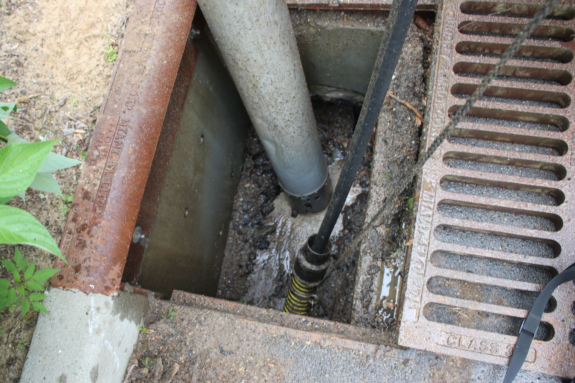 Open drain with pipes and a metal grate beside a concrete sidewalk.