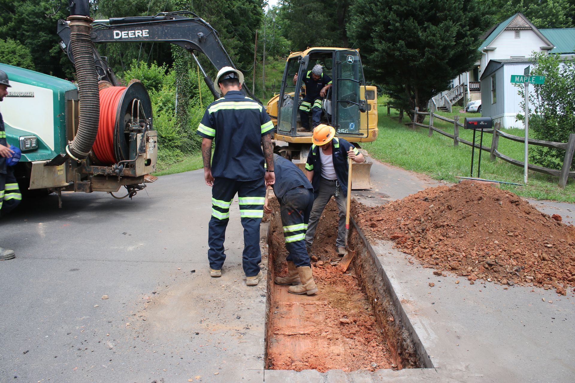 Workers digging a trench beside a road with an excavator and piles of red soil