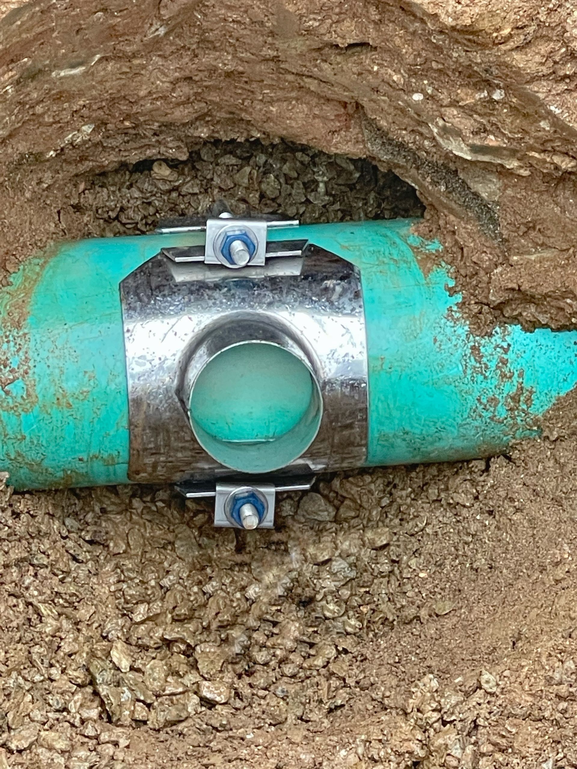 Turquoise pipe with a silver clamp and circular opening, buried in dirt under an earthen trench wall