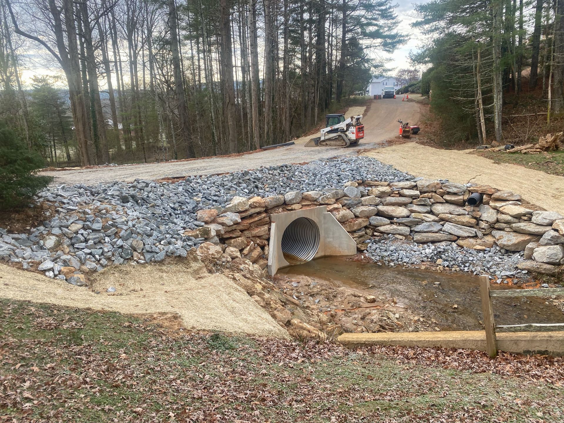 Stone-lined drainage culvert under a gravel path in a wooded area, with workers in the background