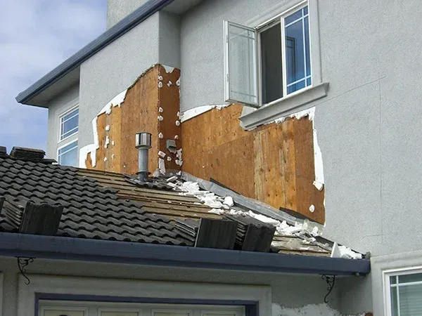 Damaged house exterior with exposed wood and collapsed roof section under a white wall