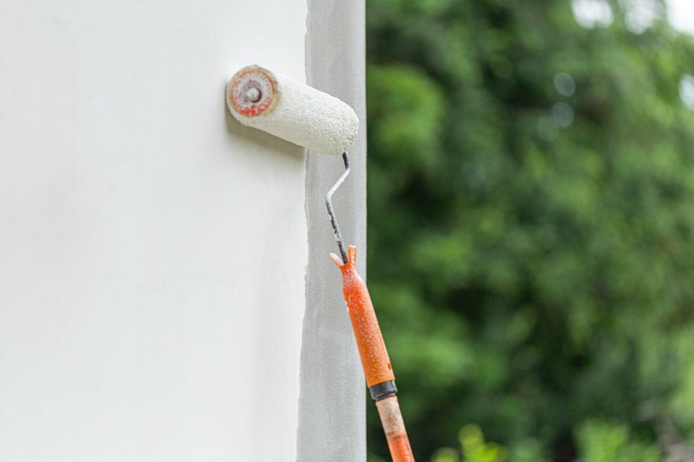 Paint roller applying white paint to an exterior wall near greenery