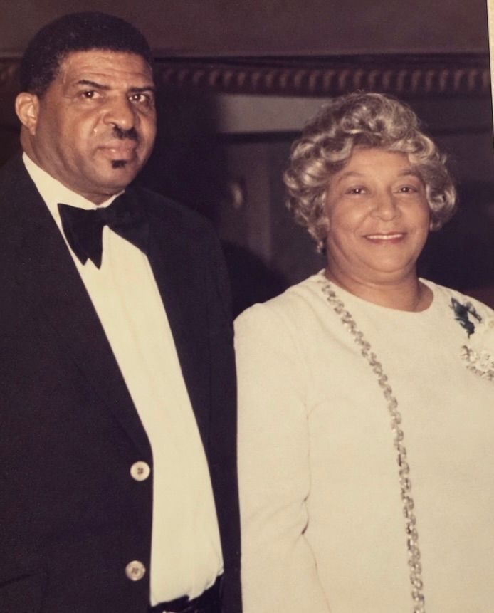 Two formally dressed adults posing together indoors, both smiling at the camera.